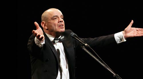 Промо-фото моноспектакля «Живой Пушкин»: артист в смокинге у микрофона разводит руки на тёмной сцене